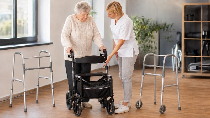 Seniorka drží madla čtyřkolového chodítka se sedátkem v obývacím pokoji