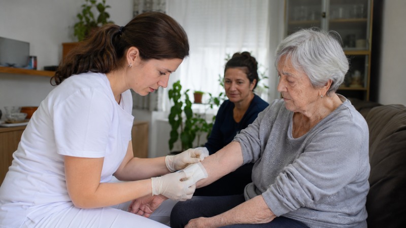 Zdravotní sestra v domácnosti převazuje seniorovi ránu na noze a měří mu krevní tlak