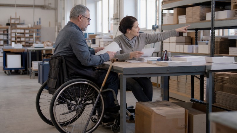 Muž na invalidním vozíku sedí u pracovního stolu ve skladu, kolegyně mu ukazuje na regál s materiálem