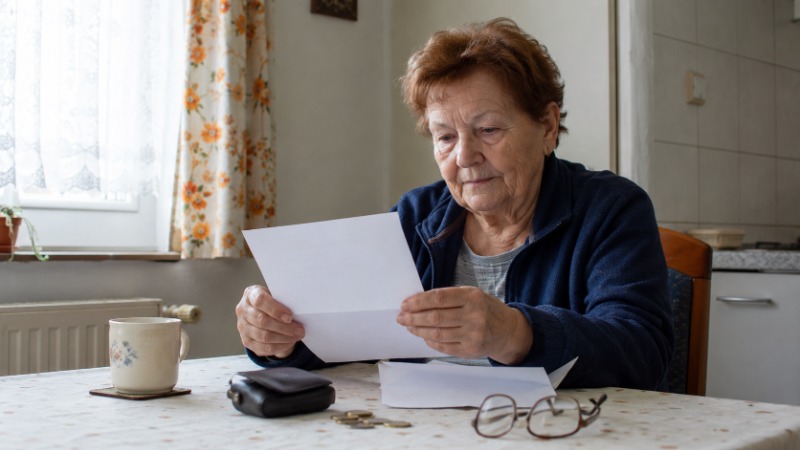 Starší žena počítá drobné mince na kuchyňském stole vedle výplatní pásky s důchodem
