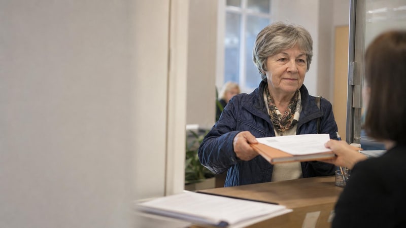 Seniorka u přepážky správy sociálního zabezpečení podává doklady k žádosti o starobní důchod