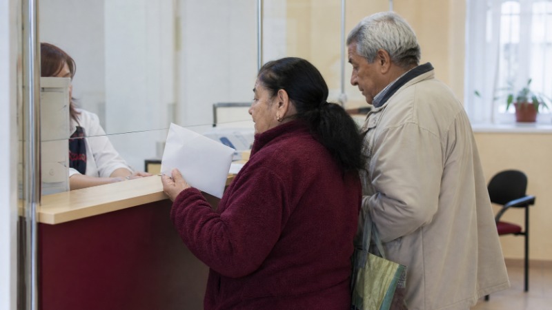 Senior otevírá u kuchyňského stolu obálku s poštovní poukázkou a dopisem od úřadu